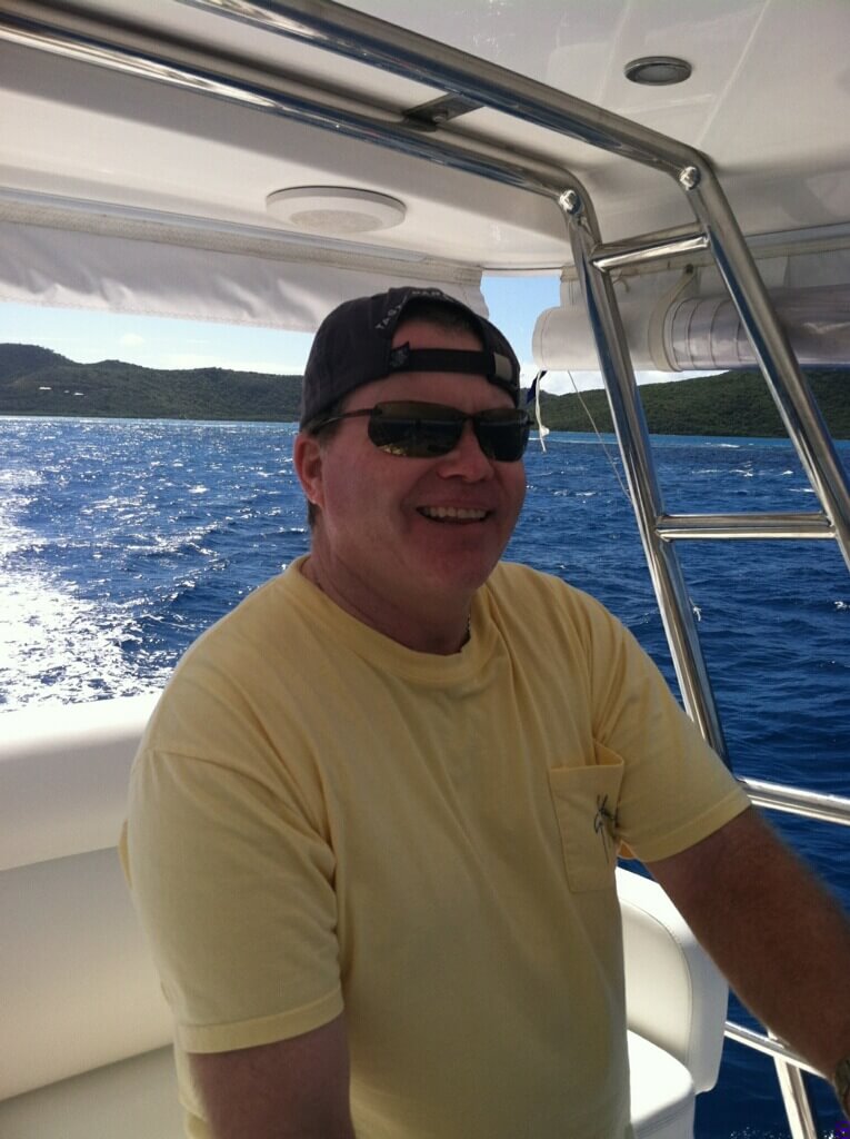 A person is smiling while sitting on a boat, wearing a yellow t-shirt and sunglasses. They have a baseball cap on backward. The background features a sparkling blue ocean and green hills in the distance, indicating a sunny day. The boat's structure includes a metal railing and a white canopy overhead.