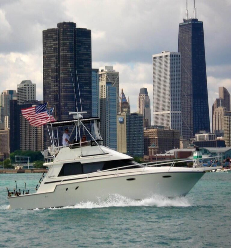 A white motorboat is cruising through the water, displaying an American flag at the stern. In the background, a skyline of tall buildings, including a prominent black skyscraper and a glass-covered structure, is visible against a cloudy sky. The water is slightly choppy, creating small waves around the boat.