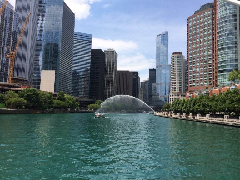 A vibrant urban scene featuring a wide waterway surrounded by tall skyscrapers. The water is a striking turquoise color, reflecting the buildings and the sky. A fountain sprays water in an arc, adding a dynamic element to the scene. On the water, a small boat is visible, navigating through the calm surface. Lush greenery lines the banks, contrasting with the modern architecture of the buildings, which include glass facades and various styles. The sky is mostly clear with a few clouds, suggesting a sunny day.